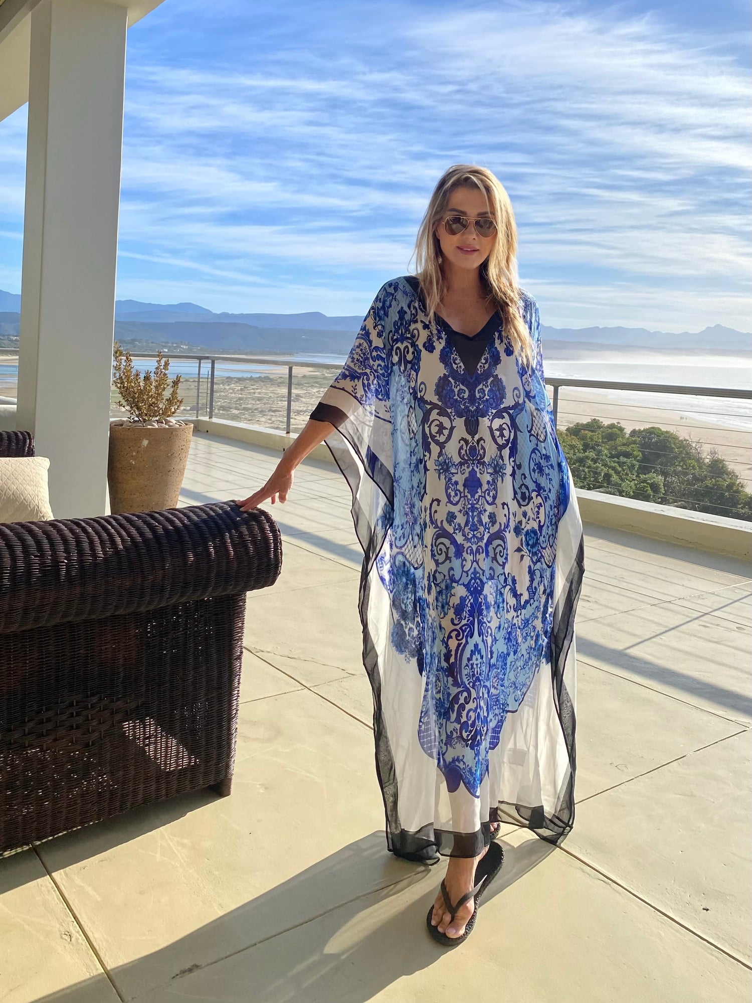 Woman in a blue and white patterned kaftan standing on a balcony with a scenic view.